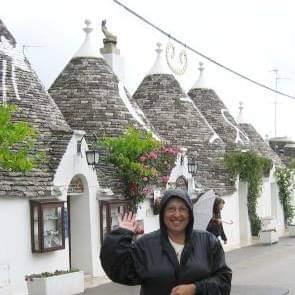 A person in a black raincoat waving with Alberobello in the backgroundAI-generated content may be incorrect.